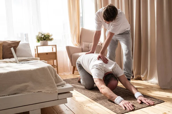 Treinador de ioga ajuda ao homem sênior a fazer ioga asana balsana corretamente em casa. — Fotografia de Stock