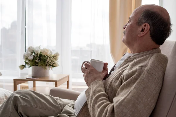 편안 한 노인 이 따뜻 한 음료를 마시며 소파에 앉아 있는 모습 — 스톡 사진