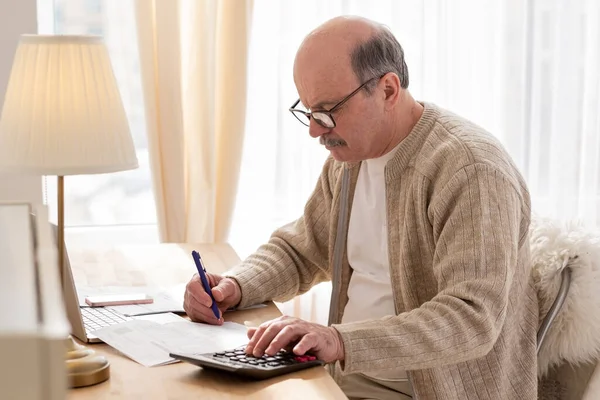 Hombre mayor sentado con papeleo y usando calculadora mientras cuenta dinero —  Fotos de Stock