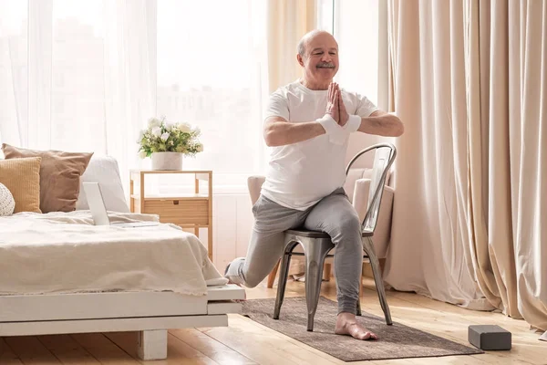 Yaşlı adam yoga yapıyor. Asana savaşçısı sandalye kullanıyor.