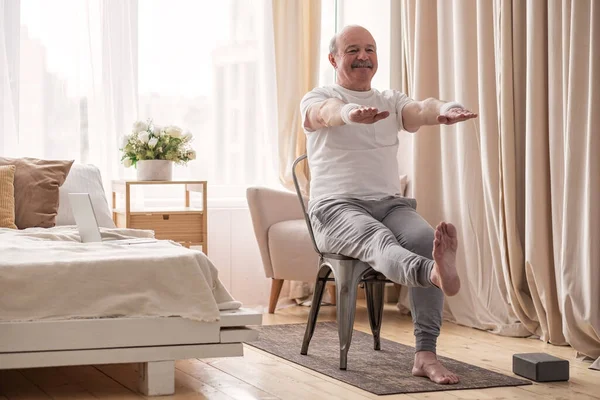 Yaşlı adam yoga yapıyor ya da sandalye üzerinde el ve ayak egzersizi yapıyor.
