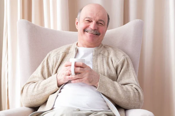 Радостный пенсионер держит в руках чашку белого кофе и смотрит в камеру — стоковое фото