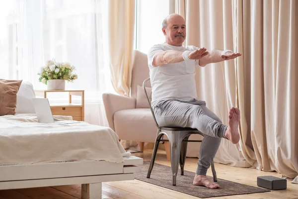 Yaşlı adam yoga yapıyor ya da sandalye üzerinde el ve ayak egzersizi yapıyor.
