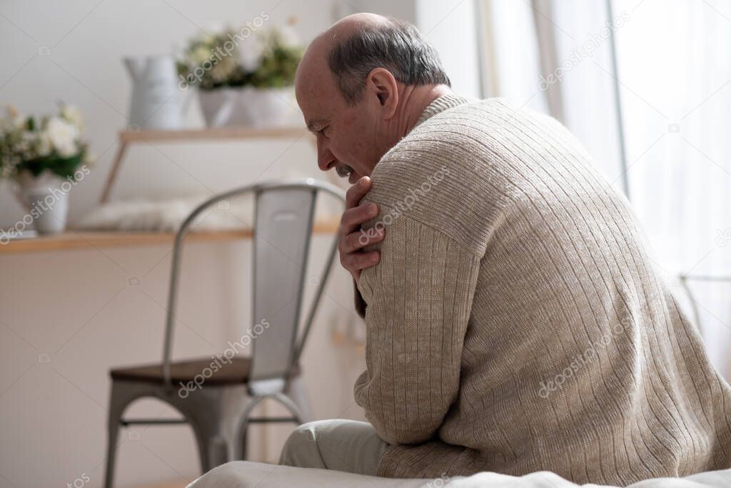 Senior man with osteoarthritis pain holding hand on his shoulder sitting at home