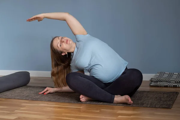 Hamile gülümseyen kadın oturuyor ve yoga yapıyor, sola eğiliyor. — Stok fotoğraf