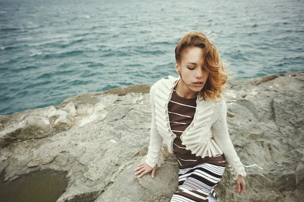 Sexy rubia vistiendo vestido con rayas y cárdigan posando sobre roca en el mar —  Fotos de Stock