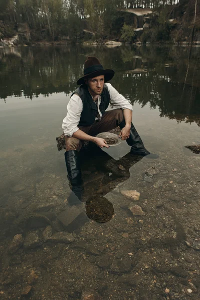 Unshaved golddigger washes gold in the lake with rocky bank — Stock Photo, Image