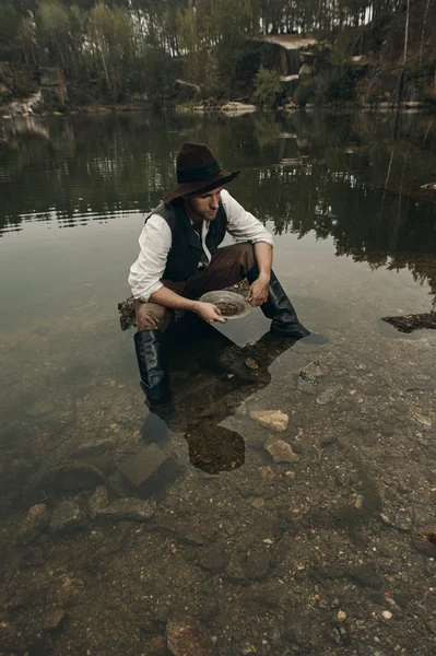 Unshaved golddigger washes gold in the lake with rocky bank — Stock Photo, Image