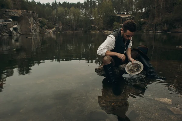 Unshaved golddigger washes gold in the lake with rocky bank — Stock Photo, Image