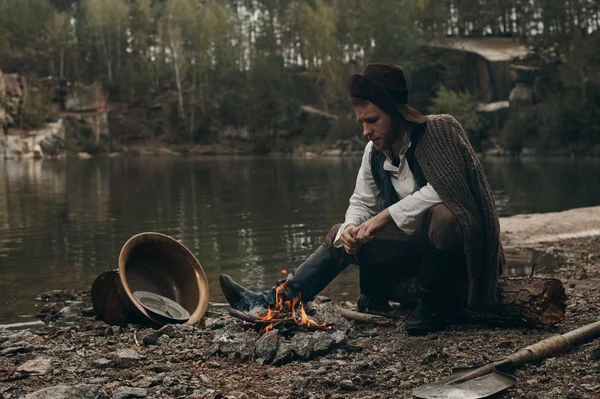 Gold digger warmin up near fire on rocky bank — Stock Photo, Image