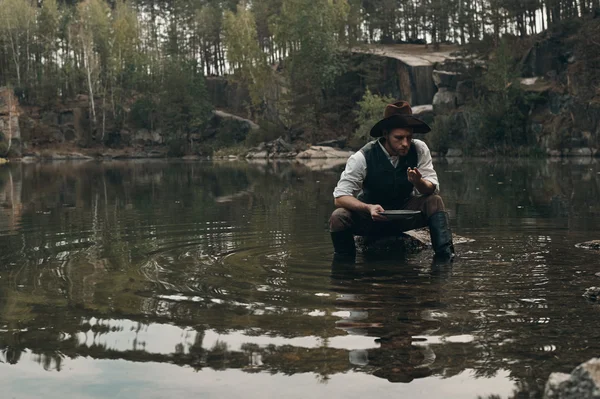 Unshaved golddigger washes gold in the lake with rocky bank — Stock Photo, Image