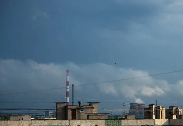 Heating and power station chimney smokestack in red and white stripes, with smoke, in an city, with rooftops, on a sunrise with dark strormy sky. Big tubes and barrels of thermal station and electro station industry. Plant pipes pollute atmosphere.