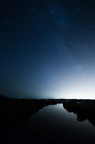 Estrellas en el cielo nocturno sobre una iluminación de fondo de la ciudad. Las luces brillantes reflejadas en la superficie plana del río . — Foto de Stock