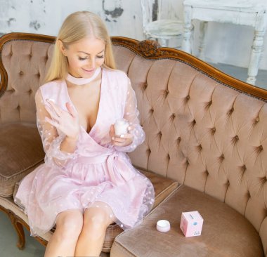  Güzel sarışın kanepede oturuyor ve elinde krema kavanozuyla pembe bir elbiseyle gülüyor.