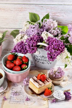 Çilekli kremalı pasta ve masada leylak çiçekleriyle duran taze meyveler.