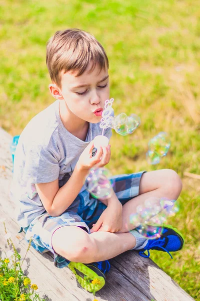 Menino sentado em um banco e soprando bolhas de sabão — Fotografia de Stock