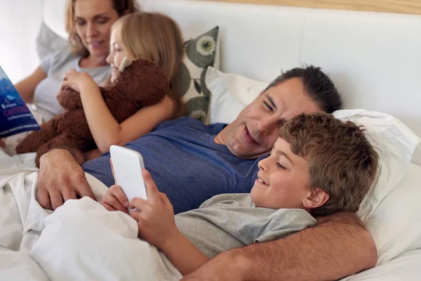 Família feliz deitada na cama com dispositivo de livro e tablet — Fotografia de Stock