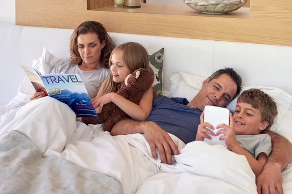 Счастливая семья лежит в постели с книгой и планшетом устройства — стоковое фото