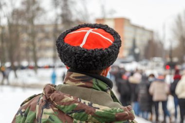 Kamuflaj ceketli ve kırmızı üzerine beyaz haçlı Kazak şapkalı bir adam bulanık insanları izliyor.