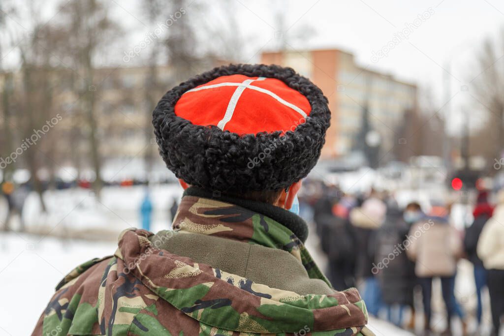 man in camouflage jacket and cossack hat with white cross on red watching blurry crowd of people