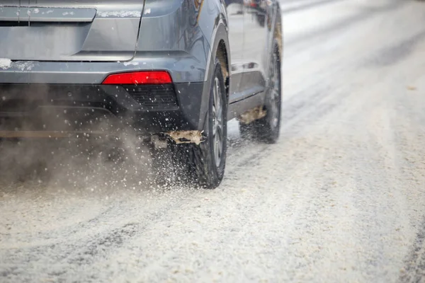 Road snow flinders flow from wheels of gray car moving fast in daylight city with selective focus.