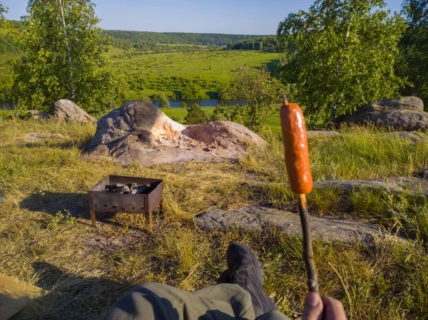 fryed sausage on a stick in front of portable brazier at wild riverside camping - gonzo view of nature recreation