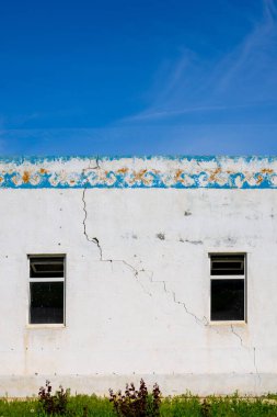Aged building stands alone in a rural area, featuring two windows and a large crack in the wall. Bleak atmosphere reflects desolation and neglect during daytime.