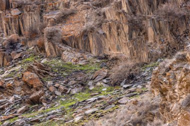 Rugged landscape features various sizes and colors of eroded sedimentary rocks, interspersed with patches of sparse vegetation. Mountainous Kyrgyzstan region captures a desolate and austere mood during daytime.
