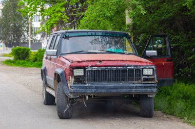 A damaged car sits abandoned on the roadside in a residential area during the daytime. The vehicle shows signs of neglect and wear, contributing to a desolate atmosphere.