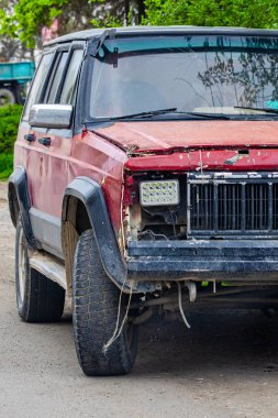 Damaged red car with significant front-end damage sits abandoned on roadside. Vegetation surrounds vehicle, conveying a sense of neglect and wear during daytime.