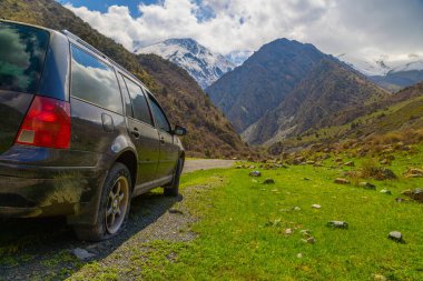 Dirty station wagon with flat tire in peaceful mountainous Kyrgyzstan region on a sunny day. Scenic outdoor setting evokes a sense of adventure and tranquility.