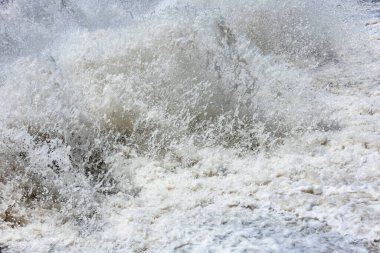 Powerful water splashes and churns in a chaotic display outdoors. The turbulent scene captures the force and energy of nature under bright daylight, evoking a sense of chaos.