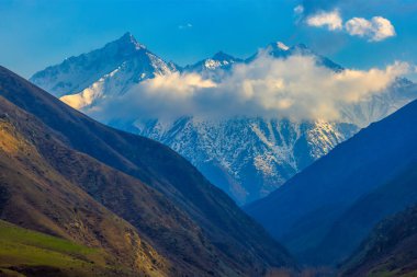A wide, scenic view of a lush valley nestled between tall mountains with snow on their peaks, under a blue sky with clouds.