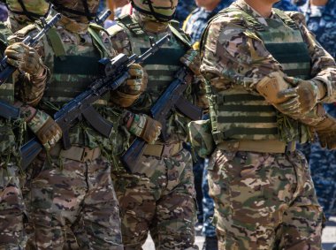 Soldiers in camouflage uniforms and tactical vests, holding assault rifles during a daytime ceremony