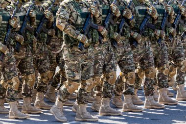 A large group of men in military uniform marches in formation on a asphalt surface under bright sunlight.