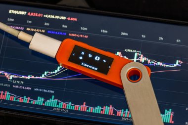 Close-up of hardware security key displaying small Ethereum logo, connected to a device. Cryptocurrency chart visible on tablet screen, highlighting technological and data-driven atmosphere.