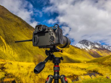 Digital camera with telephoto lens mounted on tripod focuses on majestic mountain Kyrgyzstan landscape. Serene high-altitude valley features yellow-green vegetation and small stream.