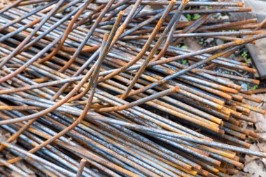 Piled metal rebar rods display rough, textured surfaces in an industrial setting. Daylight highlights rusting details, emphasizing the rugged mood of the construction site.