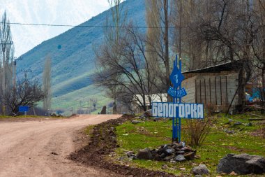 A rural scene in Belogorka, Kyrgyzstan, featuring a bright blue sign marking the village name against a mountainous landscape. The sign stands beside a dirt road, amidst sparse vegetation and trees, under a cloudy sky - April 4, 2024