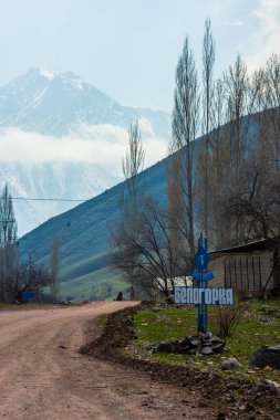 A scenic view of Belogorka, Kyrgyzstan, featuring the village sign set against a backdrop of mountains and a cloudy sky. The sign is positioned along a dirt road, amidst sparse vegetation and trees, capturing the rural essence of the area - April 4