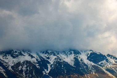 Snow-covered mountains rise majestically, partially hidden by dramatic clouds during daytime. The scene evokes a sense of mystery and serenity in a cold outdoor setting.