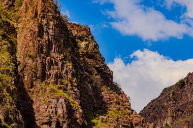 Vast mountainous region showcases rugged sedimentary rock formations and steep cliff faces. Lush vegetation contrasts with rocky textures, creating a serene outdoor landscape.