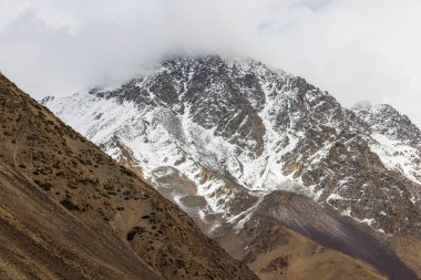 Snow-covered rocky mountains rise majestically in a serene mountainous Kyrgyzstan region. Dramatic clouds create a cold atmosphere, showcasing the beauty of nature during daytime.