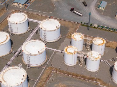 Aerial view reveals multiple white cylindrical storage tanks connected by pipes and walkways. Tanker truck in background highlights industrial setting during daytime.