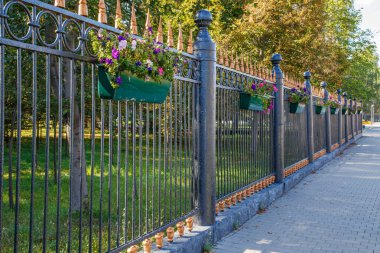 Ornamental iron fence adorned with vibrant flower planters creates a serene atmosphere in an urban park. Daytime setting enhances the peaceful and pleasant mood, showcasing natures beauty.