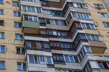 Çok sayıda pencere ve balkonun bulunduğu bir şehir binasının dış görünüşü, bazıları yıpranma ve bakımsızlık belirtileri gösteriyor. Sahne, gündüz vakti kasvetli ve yaşlanmış bir atmosferi taşır..