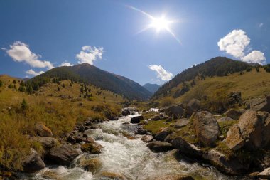 Rocky Dağı nehri bir Zhylamysh Boğazı 'ndan taşar. Çimlerle kaplı güneşli yamaçlar ve dağınık ardıç ağaçları bulutlarla dolu açık mavi bir gökyüzünün altında yükseliyor..
