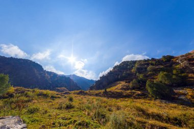 Geniş bir açı dağ vadisine bakıyor. Çimenli yamaçlar ve her yer yeşil ağaçlarla kaplı. Güneş parlak bir şekilde, açık mavi gökyüzünde ince bulutlar tarafından kısmen gizleniyor. Gölgeler yeryüzüne düşer.