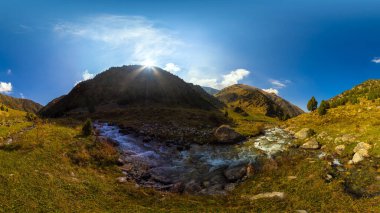 Geniş bir manzara, iki temiz akarsuyun birleştiği ve yokuş aşağı aktığı bir dağ vadisini gösteriyor. Vadi yamaçları yeşil ve sarı otlarla kaplıdır. Dağılmış kayalar ve birkaç yeşil ağaç vardır. Güneş dağın birinin arkasından ışıl ışıl parlıyor.