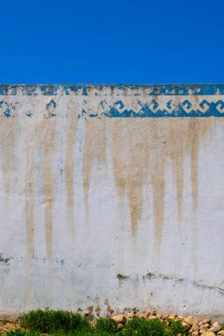 Açık gri duvarlı, bronzlaşmış ve soluk mavi desenli sınır açık havada güneşli gökyüzünün altında duruyor. Kırsal cazibe, sahnenin zemin örtüsüyle çevrelenmiş karakterini artırır..